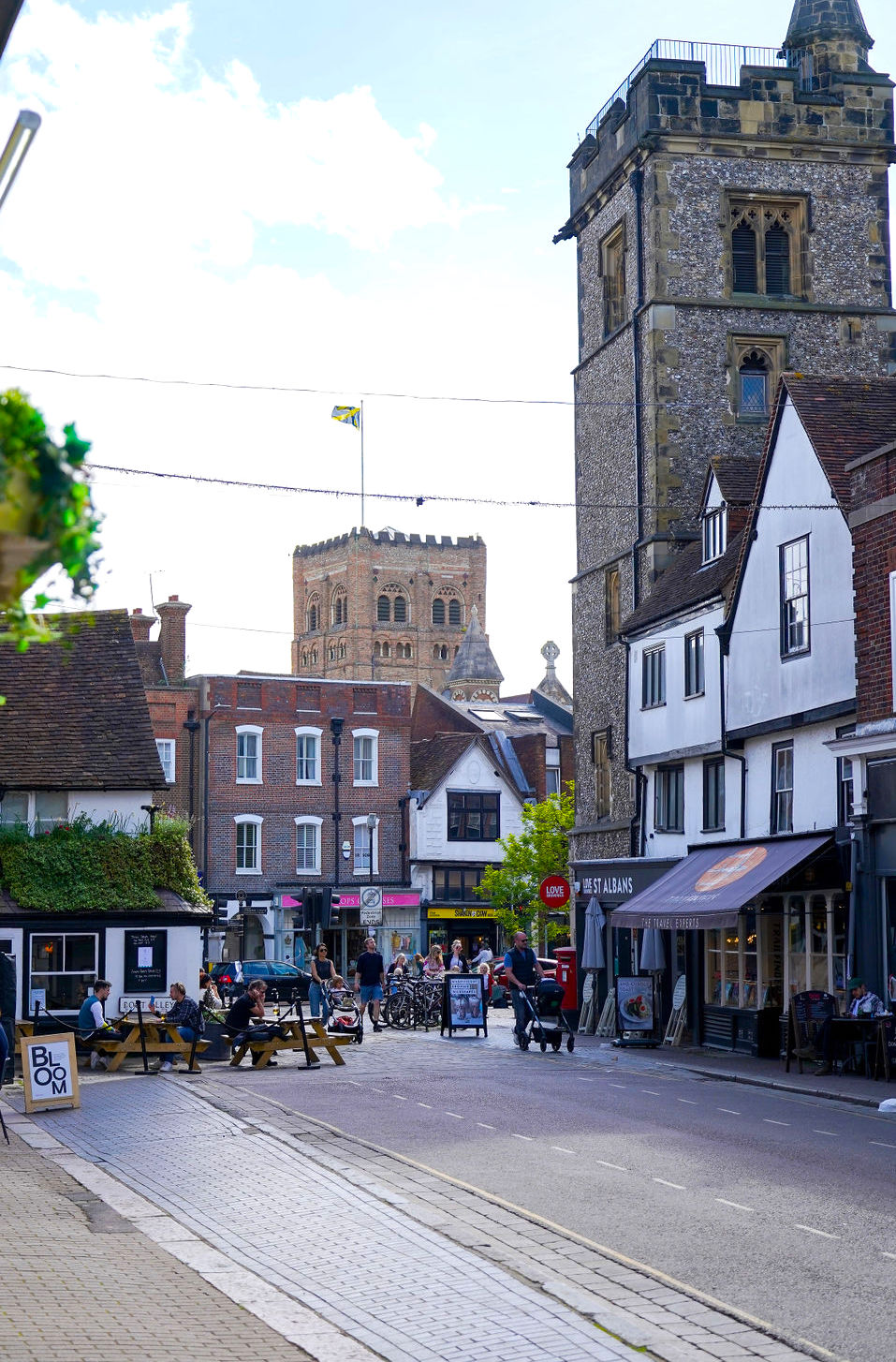 Street scene in St Albans with people sitting outside The Boot pub, historic buildings lining the road, and the cathedral tower rising in the background.